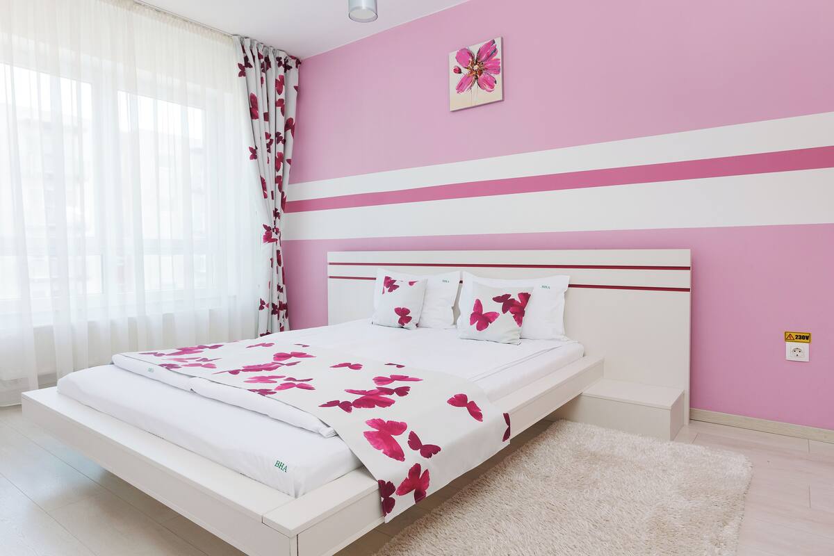A bedroom with a bed, white bedding, a pink wall, and a window with sheer curtains.