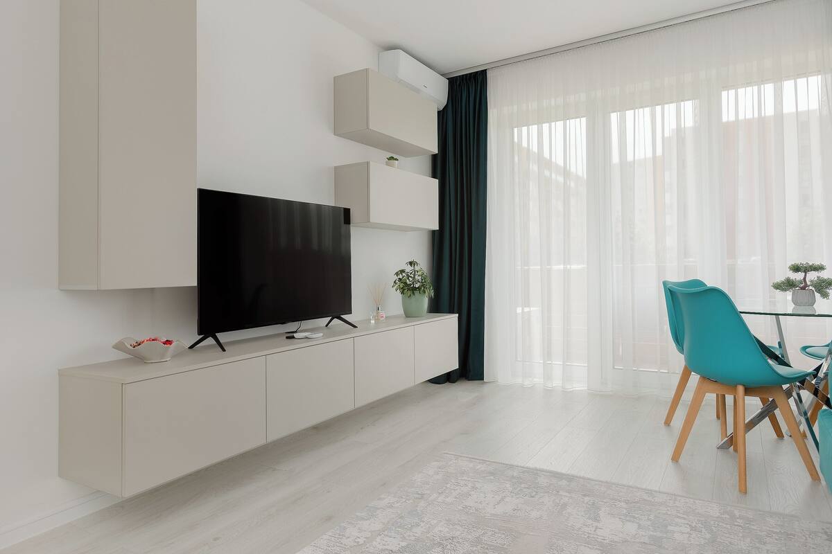 A modern living room with a flat-screen TV, a white cabinet, teal chairs, and a glass dining table.