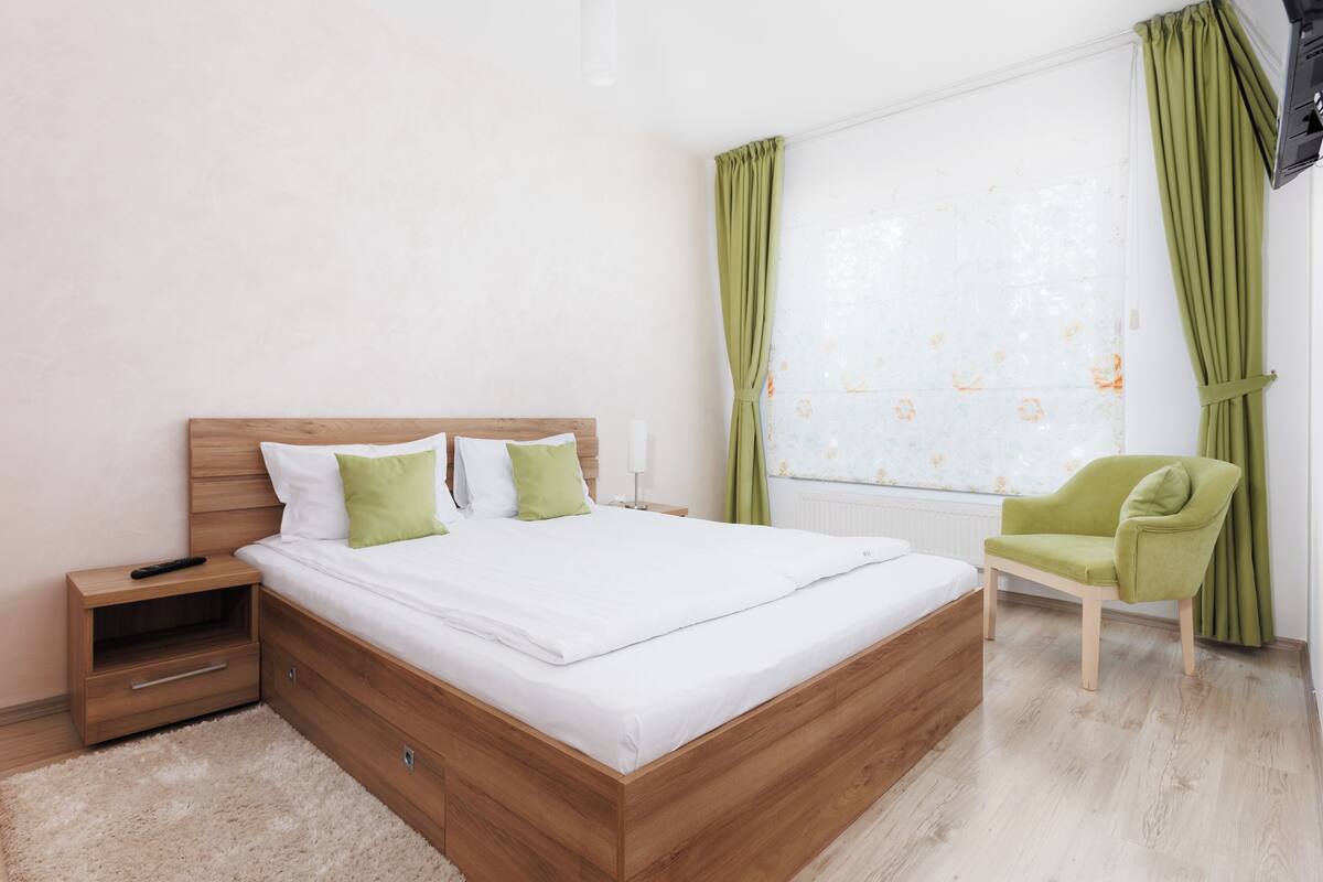 A bedroom with a wooden bed, green pillows, a nightstand, a green chair, and a window with curtains.