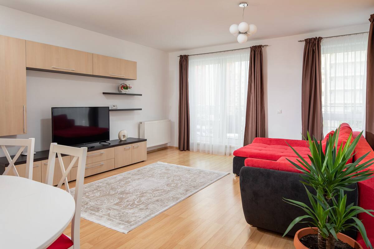 A modern living room with a flat-screen TV, a dining table with chairs, a red sofa, a potted plant, and large windows with curtains.