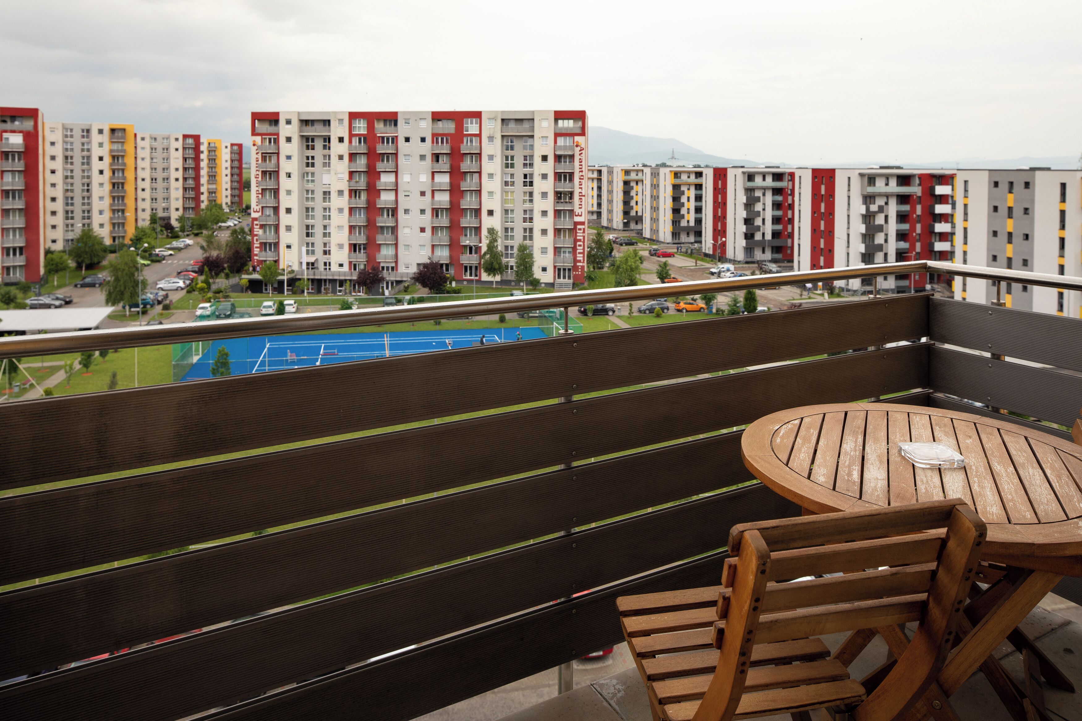 A balcony with a table and chairs, overlooking a tennis court and residential buildings.