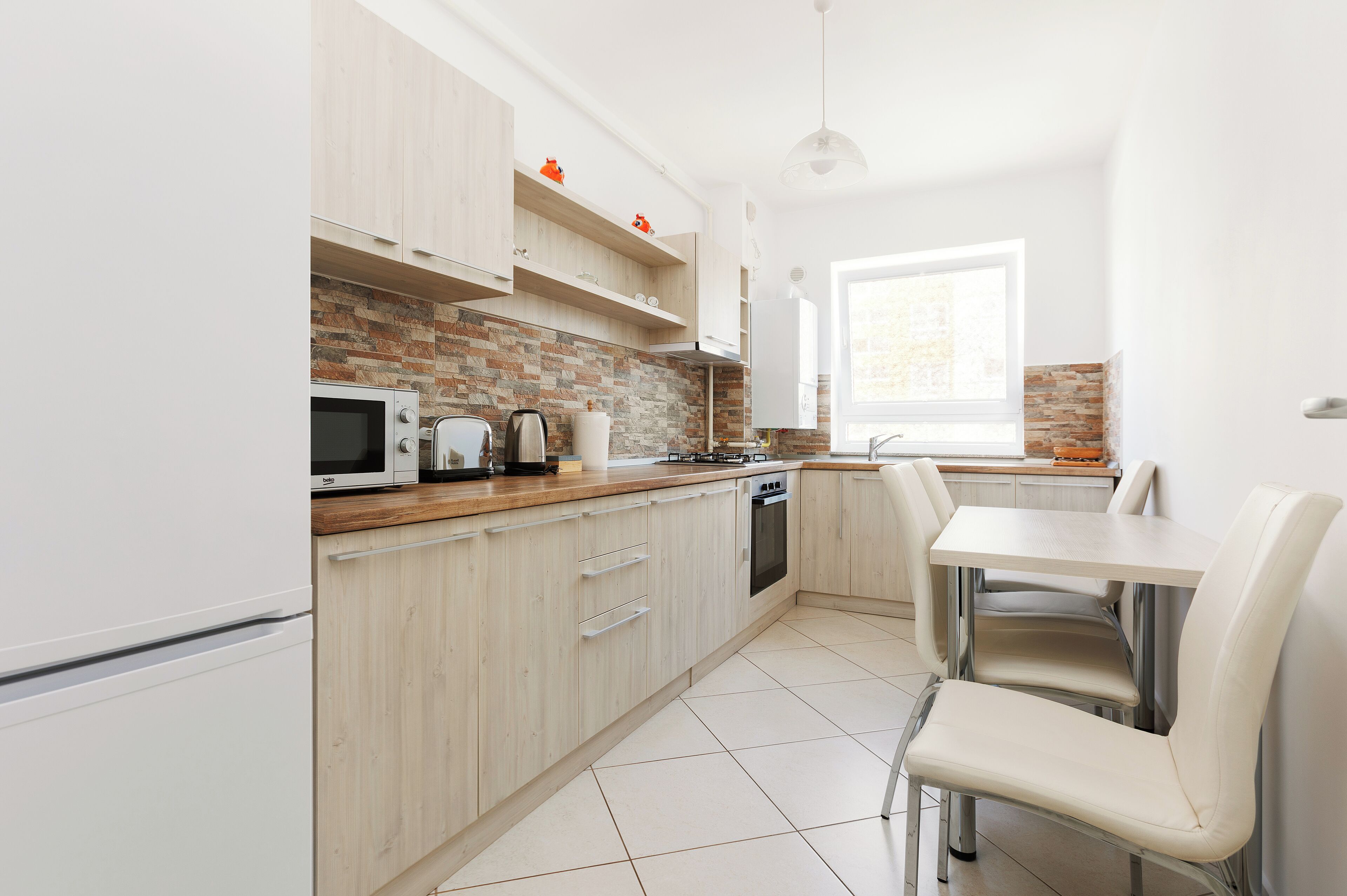 Une cuisine moderne avec des meubles de couleur bois clair, un mur de fond en briques, un four à micro-ondes, une bouilloire et un coin repas comprenant une table et des chaises.