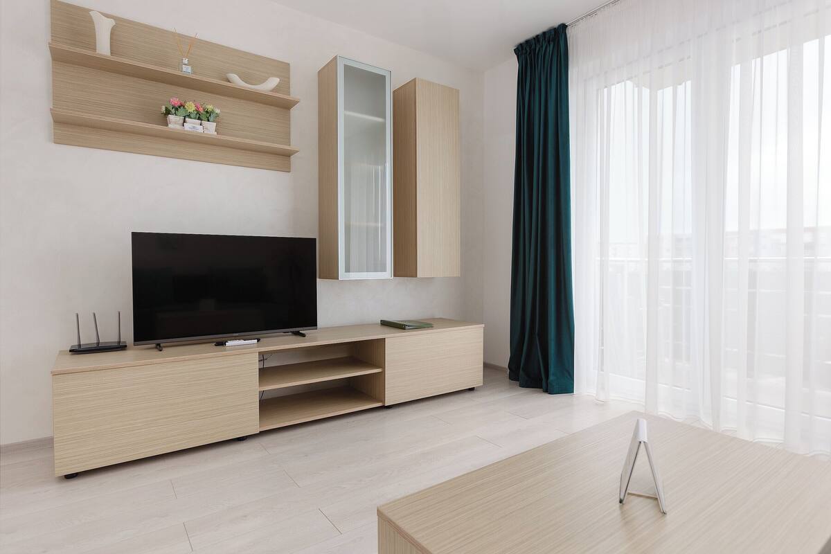 A modern living room with a flat-screen TV, wooden entertainment unit, and a large window with sheer curtains.