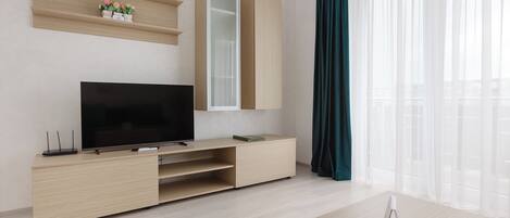 A modern living room with a flat-screen TV, wooden entertainment unit, and a large window with sheer curtains.