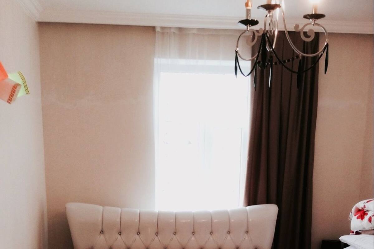 A bedroom with a tufted headboard, a bed with a beige bedspread, two pillows, a teddy bear, and a chandelier.