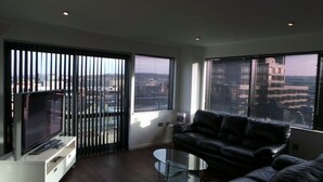 A modern living room with a black leather sofa, a glass coffee table, and a flat-screen TV on a white stand. Large windows with blinds allow natural light and views of the cityscape. - City Crash Pad - West Street Apartments (Sheffield)