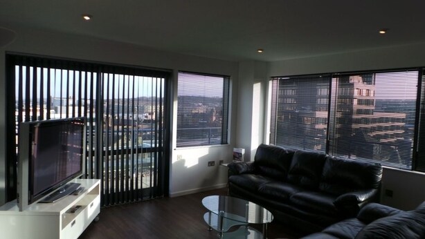 A modern living room with a black leather sofa, a glass coffee table, and a flat-screen TV on a white stand. Large windows with blinds allow natural light and views of the cityscape.