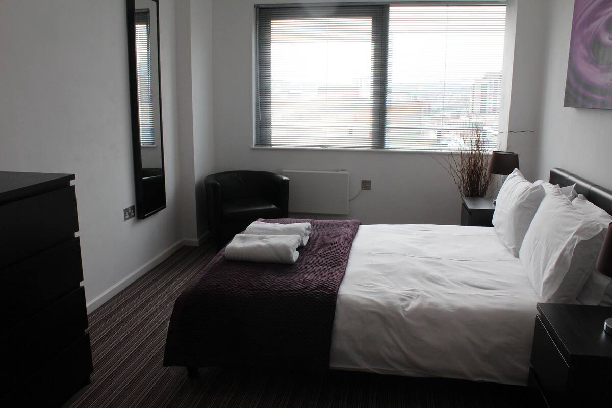 Une chambre d’hôtel avec un lit, une chaise, une commode et une fenêtre avec des stores.