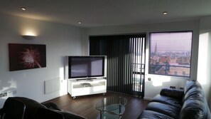 Una sala de estar moderna con un televisor de pantalla plana sobre un mueble blanco, una mesa de centro de cristal, un sofá de cuero y un ventanal con persianas.