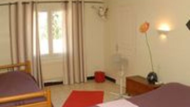 A hotel room with two beds, a wooden headboard, a window with curtains, a bedside table, and a lamp. - Hôtel les Terrasses du Rove (Le Rove)