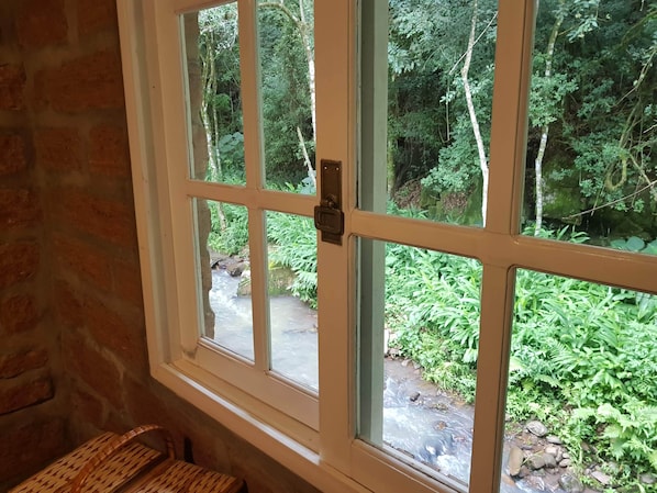 A room with a window overlooking a natural landscape with a stream and dense foliage. - Villa Italia Hospedagem (Gramado)