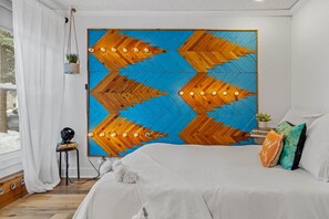 Une chambre avec un grand lit, un luminaire fixé au mur, une petite table avec une plante et une fenêtre avec des rideaux.