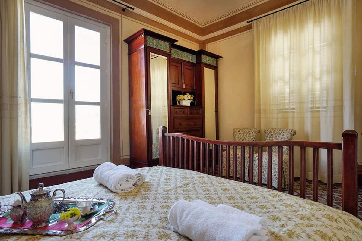 Une chambre d’hôtel avec un grand lit, une commode en bois, une chaise et une fenêtre avec des rideaux.