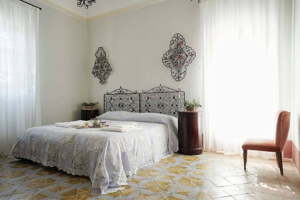 Une chambre avec une couvre-lit à motifs, une tête de lit décorative, une plante en pot, une table de chevet en bois et une chaise.