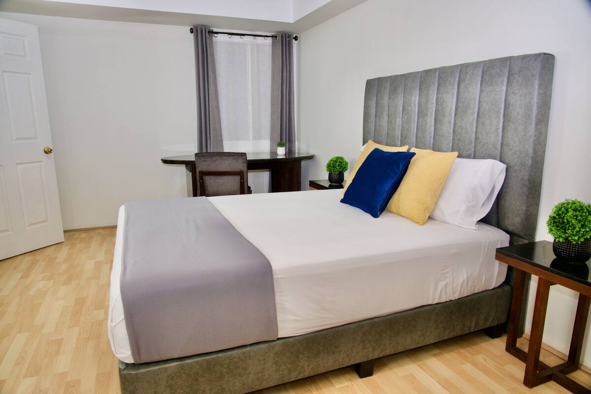 A neatly made bed with a grey headboard, a bedside table with a plant, and a chair in a room with wooden flooring and a white door.