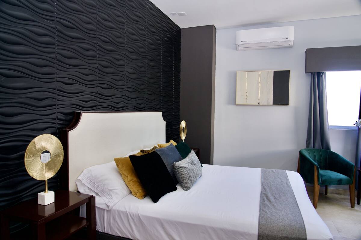 A modern hotel room with a large bed, a bedside table with a decorative lamp, a wall-mounted air conditioner, a window with curtains, and a green armchair.
