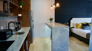 A modern kitchen with granite countertops, a built-in oven, and a refrigerator. There is a dining area with a table and chairs, and a bed with a dark headboard and multiple pillows. - Rio Suites (Tijuana)