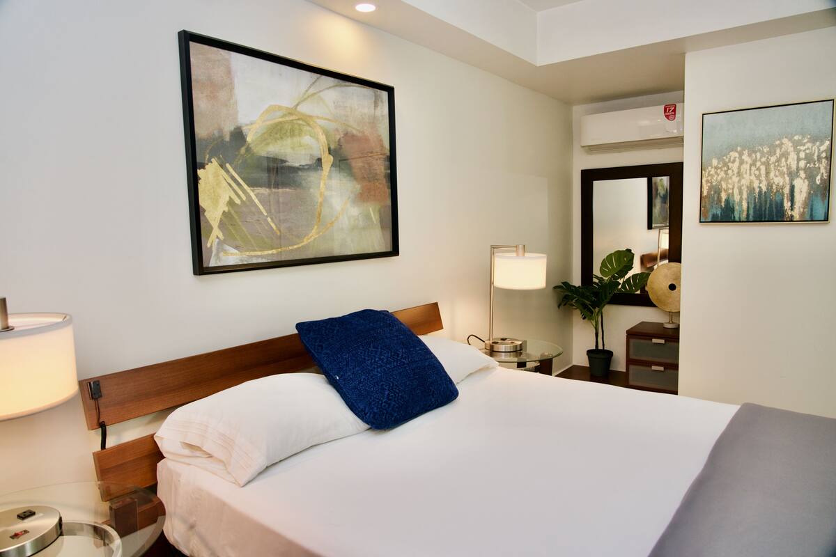 A hotel room with a large bed, a nightstand with a lamp, a potted plant, and artwork on the walls.