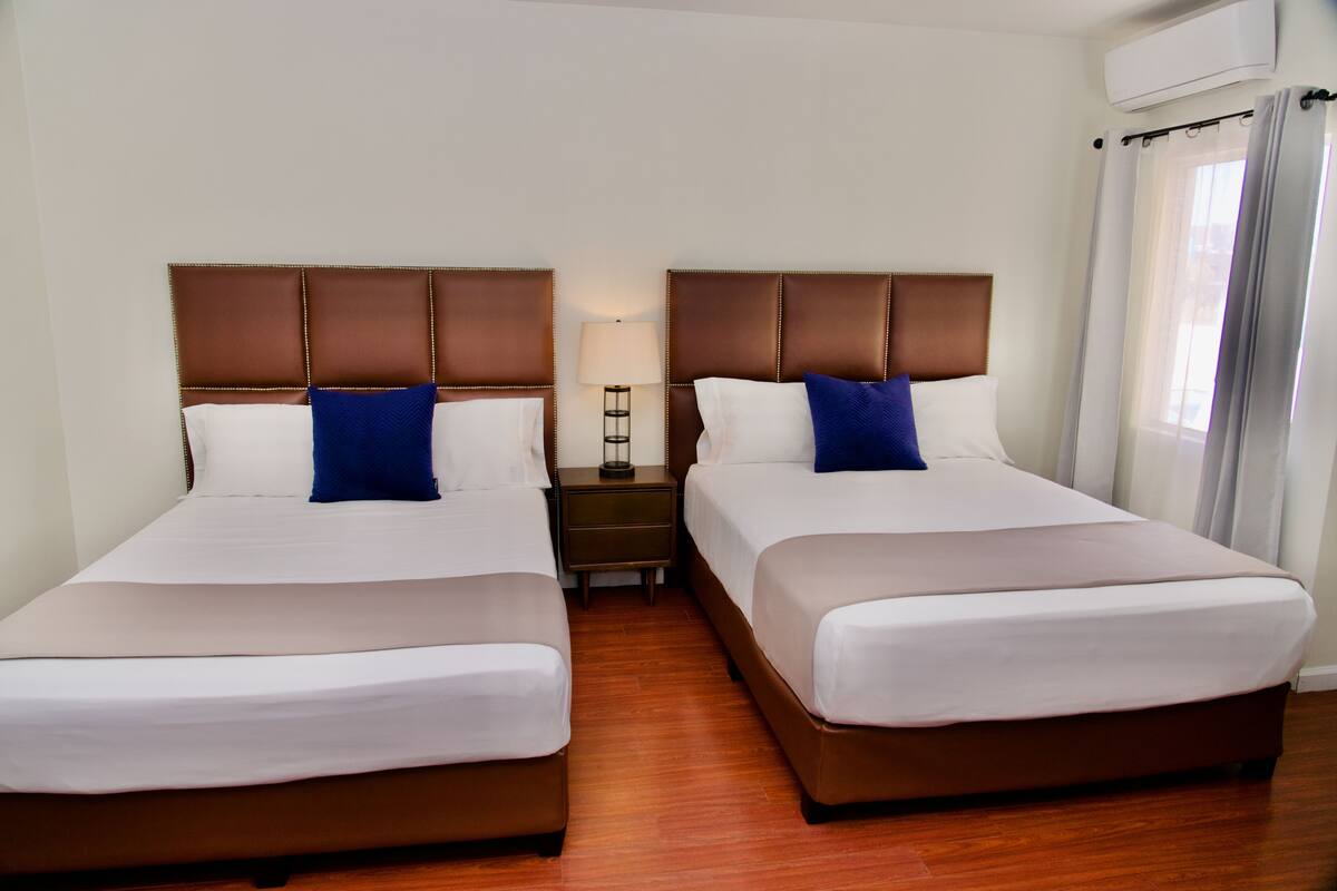 A hotel room with two beds, a nightstand, a lamp, and an air conditioning unit.