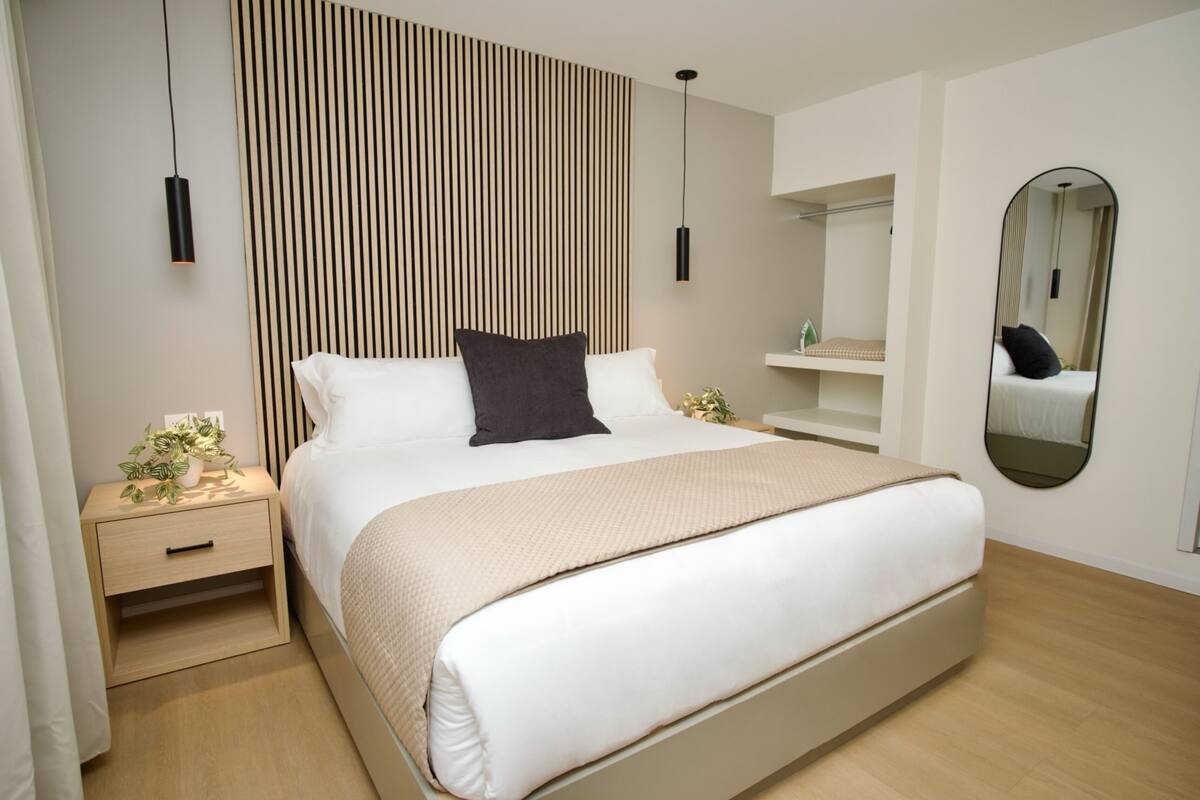 A modern bedroom with a large bed, bedside tables, a mirror, and a vertical wooden panel wall.
