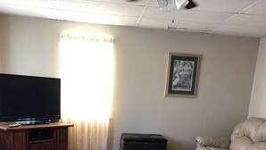 Ein Wohnzimmer mit Fernseher auf einem Holzständer, einem großen, bunten Teppich, einer Ledersofa, einem beigen Sessel und einem gerahmten Bild an der Wand.