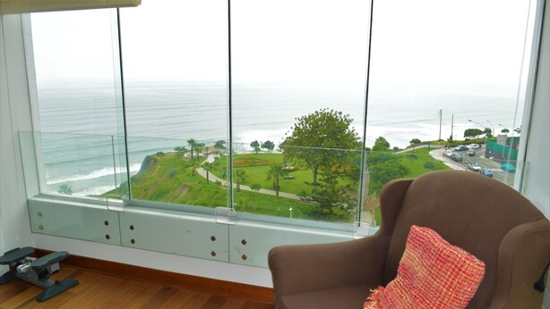 Una habitación con un ventanal grande que da a un paisaje costero, una silla marrón con cojín rojo y una pequeña mesa con una silla.