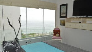 Un dormitorio con un ventanal grande con vistas al océano, una cama con almohadones estampados, una silla con cojín y un televisor en la pared.