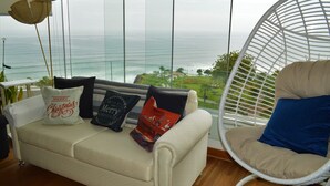 Una sala de estar con vista al océano, un sofá blanco con almohadones decorativos y una silla de mimbre blanca.