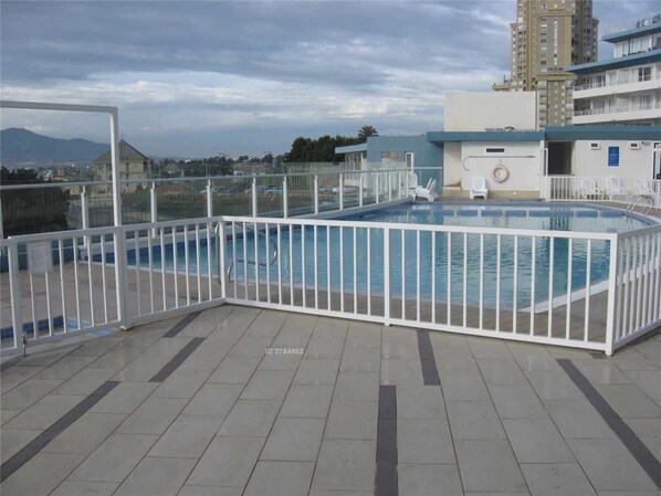 Une piscine sur le toit, entourée d’une clôture blanche, avec un sol carrelé et une vue sur les bâtiments et les montagnes en arrière-plan.