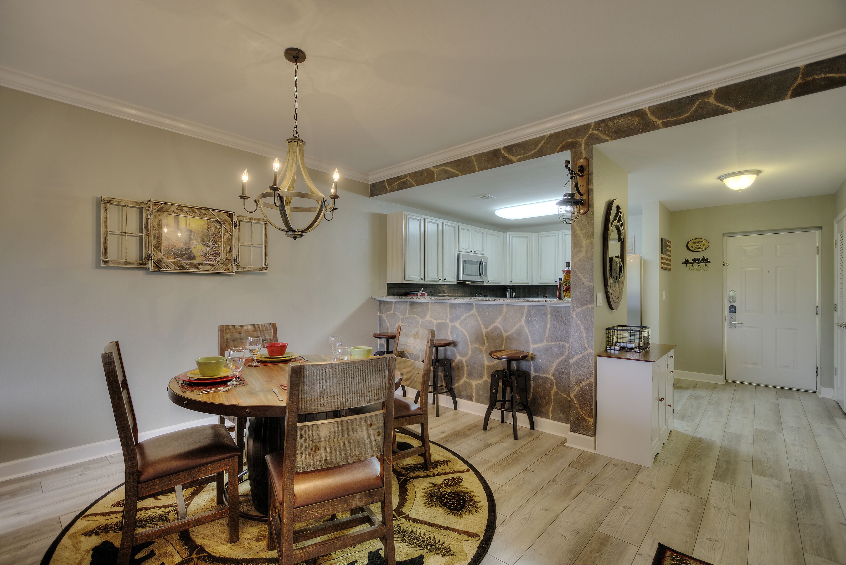 A dining area with a round table, chairs, and a chandelier. There is a kitchen area with cabinets and a microwave in the background.
