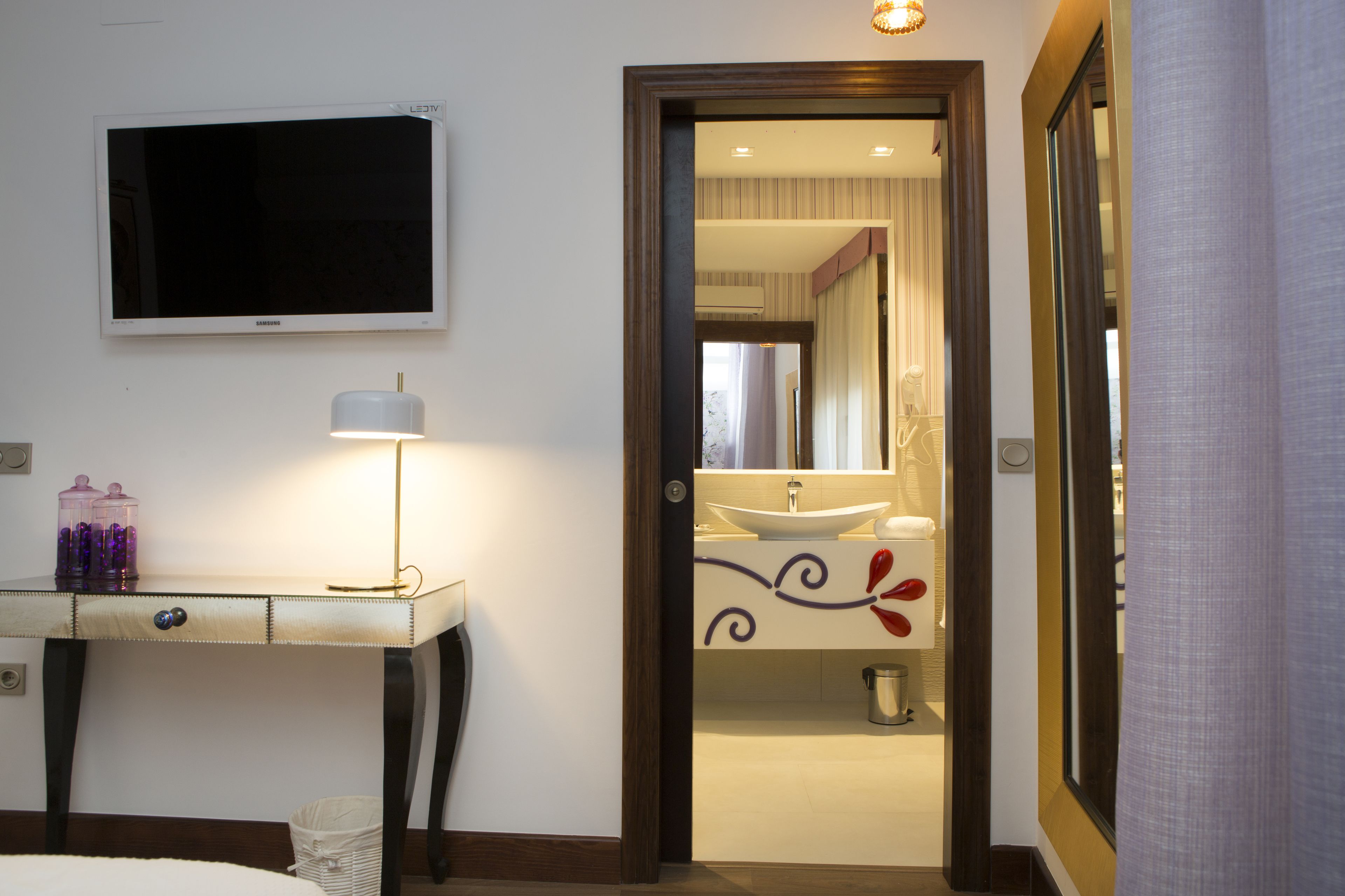 Habitación de hotel con televisión montada en la pared, una mesita de noche con lámpara, y un ba ño visible a través de una puerta abierta.