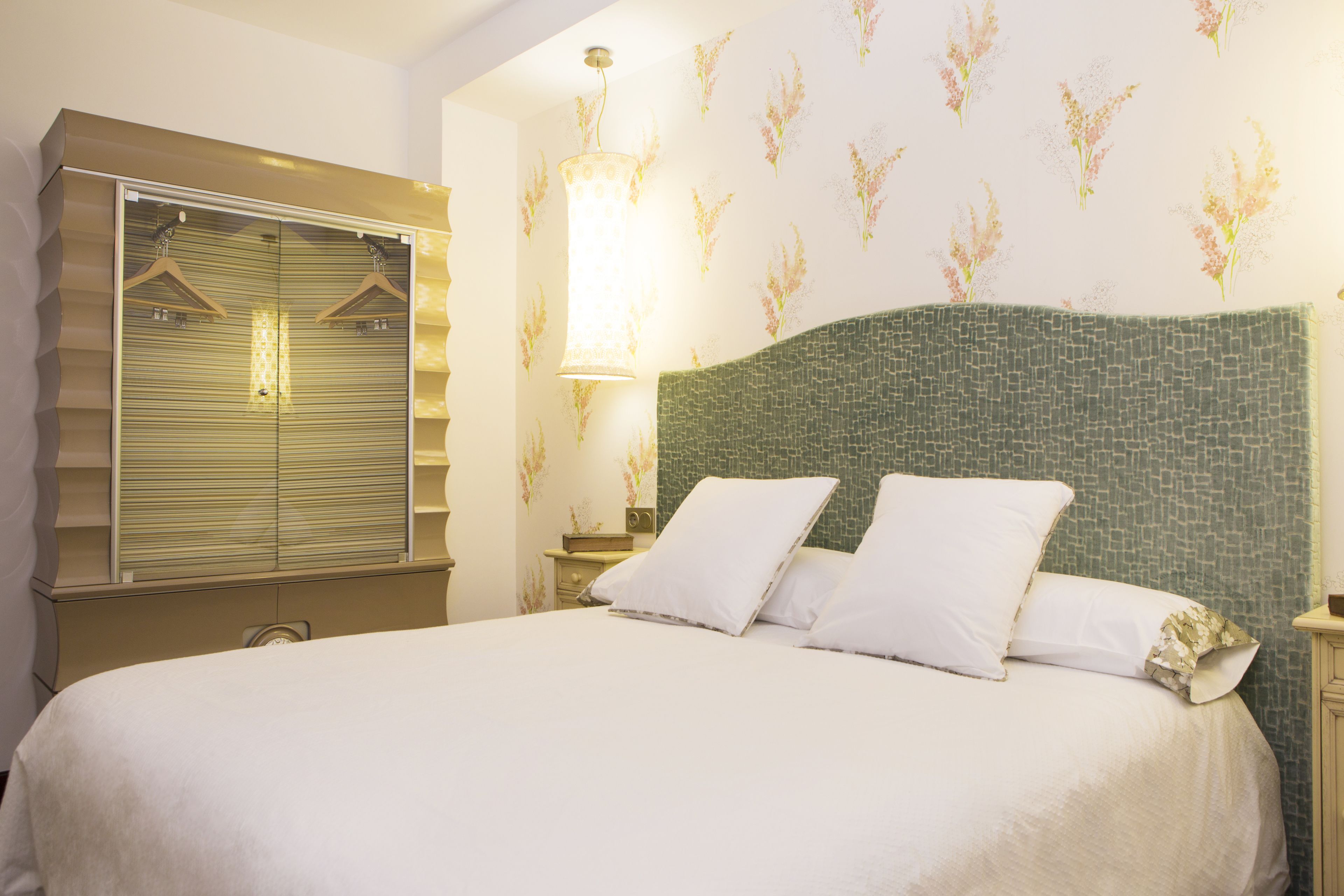 A neatly made bed with a green headboard, a large mirror with a wooden frame, and a wall with floral wallpaper.