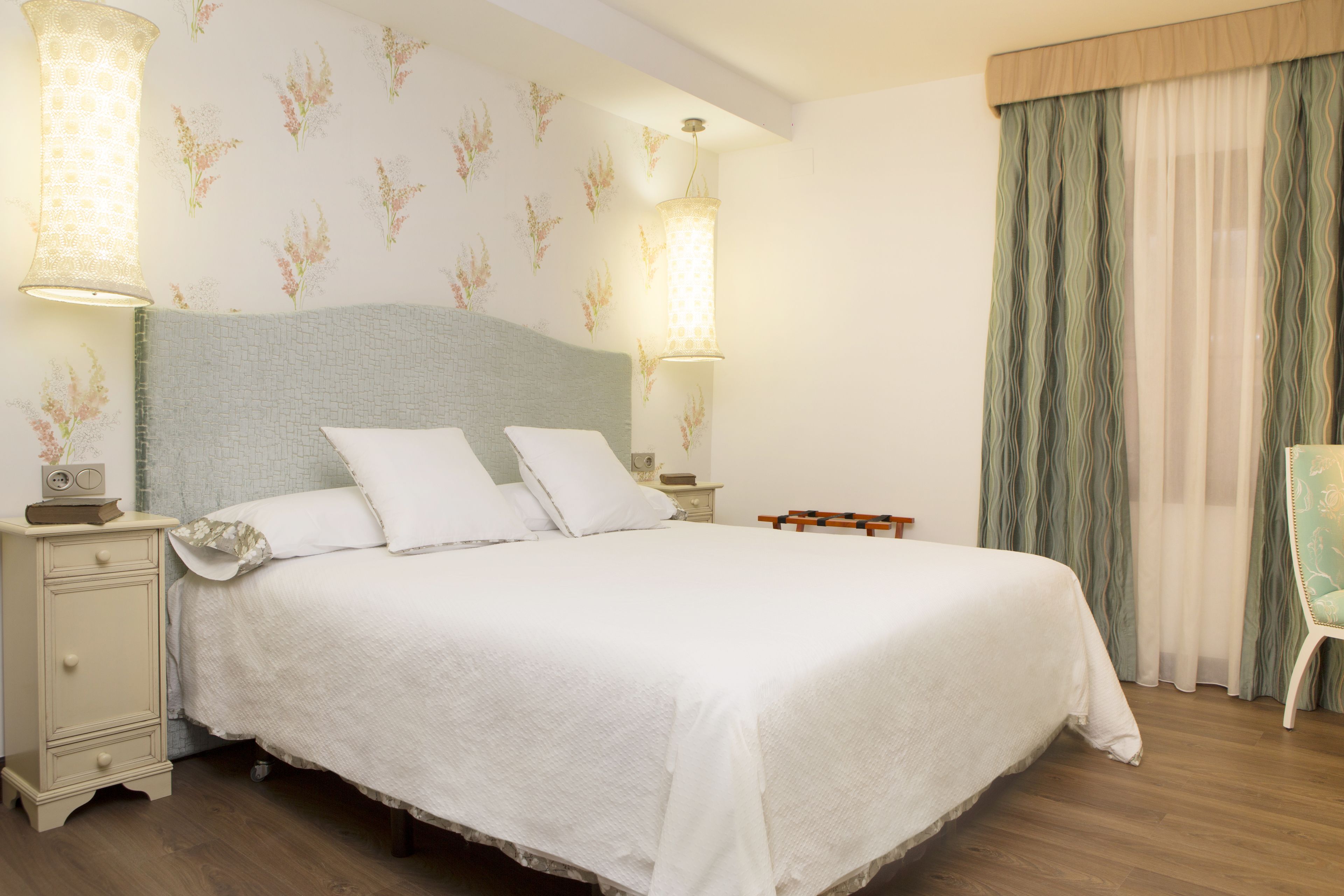 Een netjes opgemaakt bed met witte lakens en kussens, twee bedlampen, een nachtkastje met een radio, en een stoel met een gestreept kussen.