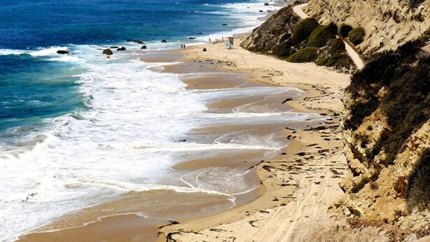 A coastal beach with a clear view of the ocean, sandy shore, and a rocky cliff.