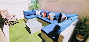 A sectional sofa with blue cushions, a wooden chair, a small white ottoman, and a potted plant.