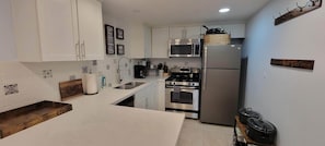 A compact kitchen with white cabinets, a stainless steel oven, a microwave, and a refrigerator. There are decorative tiles on the backsplash and a wooden shelf.