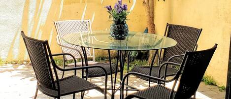 Un patio con una mesa de vidrio y sillas de mimbre, rodeado por un muro amarillo y follaje.