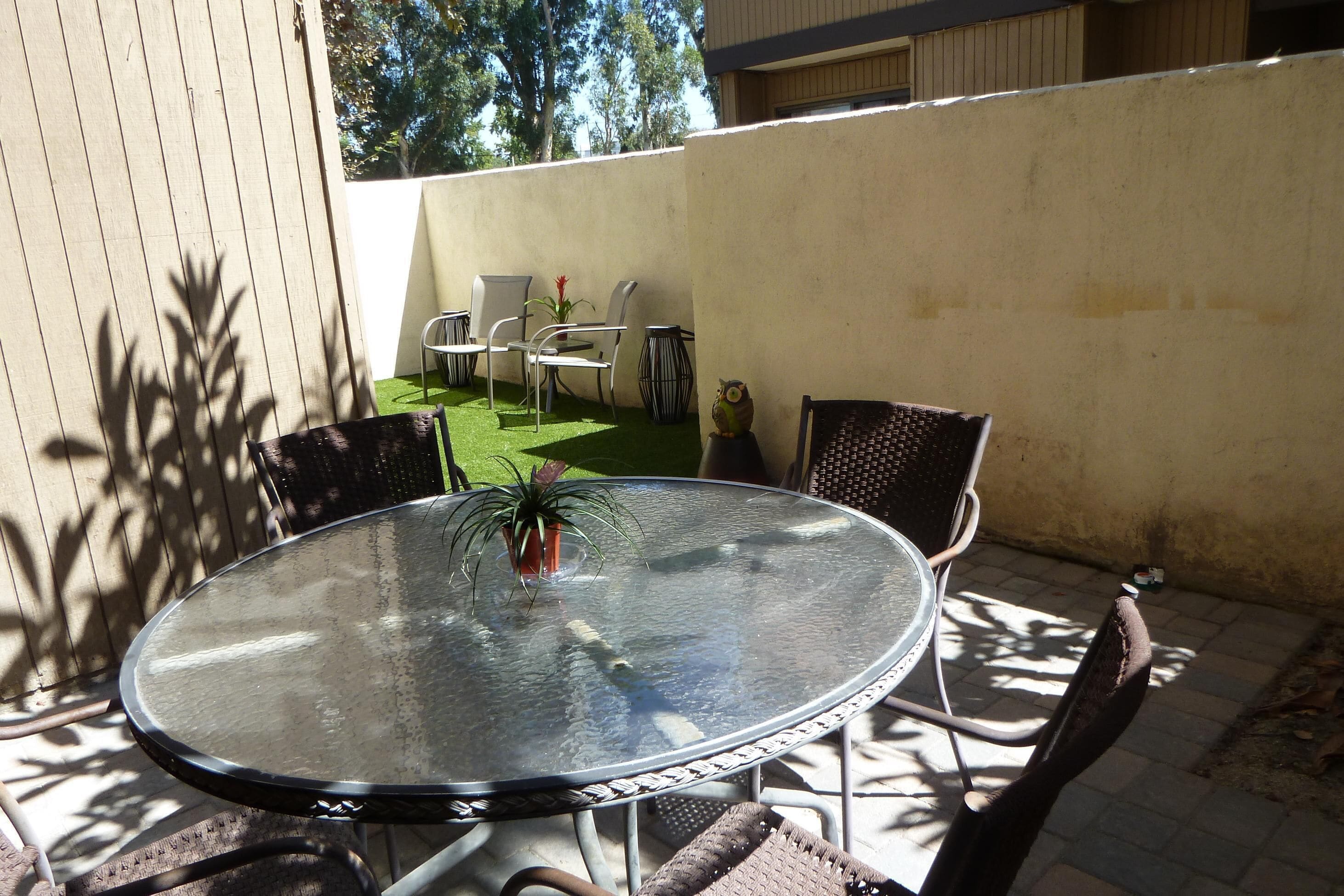 A glass-top outdoor table with metal chairs, surrounded by a small green lawn and a white wall.