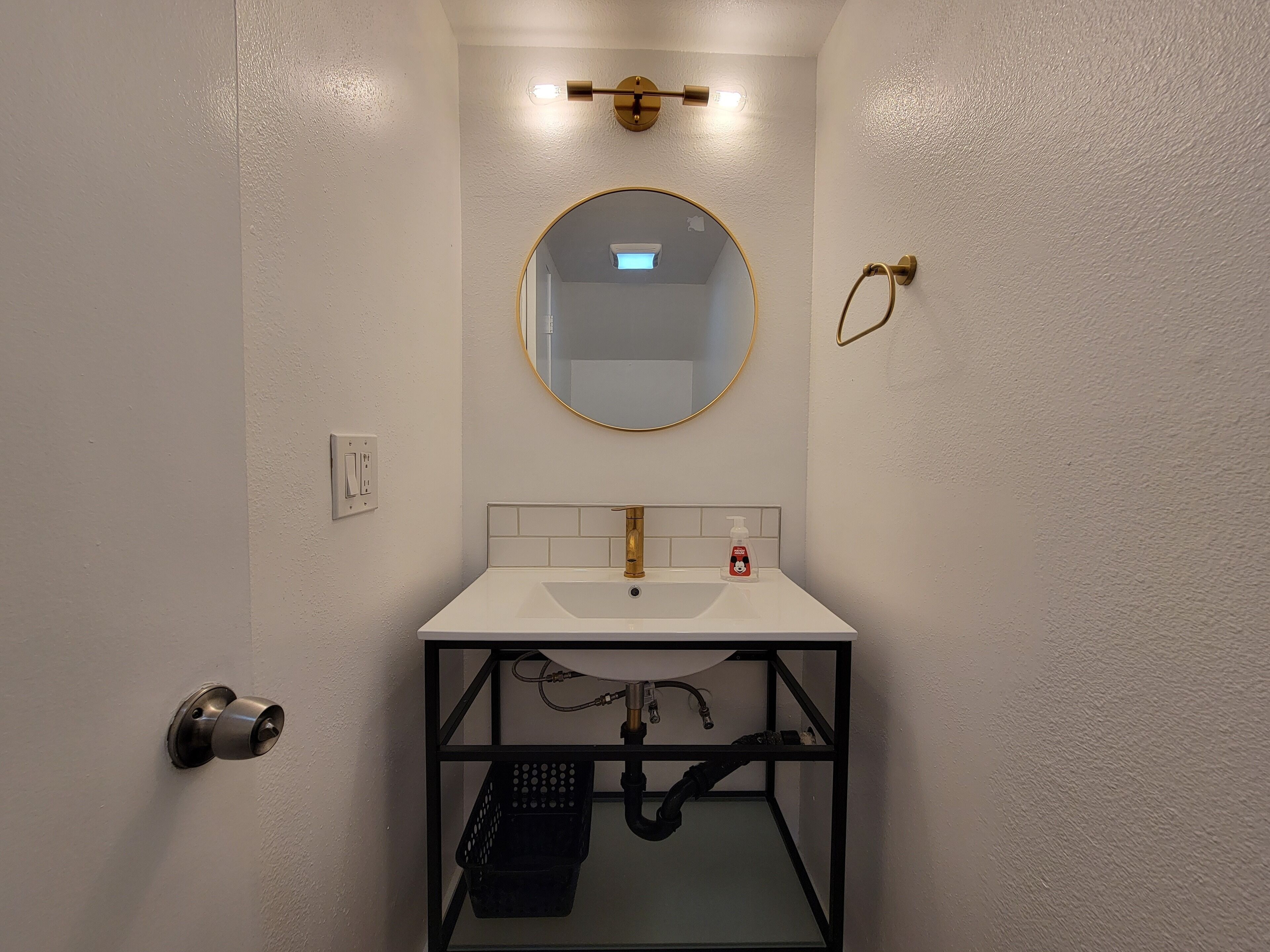Un baño con lavamanos blanco, accesorios dorados, un espejo redondo y un estante metálico negro.