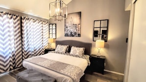 A bedroom with a large bed, two bedside tables with lamps, a headboard, and a cityscape painting on the wall.