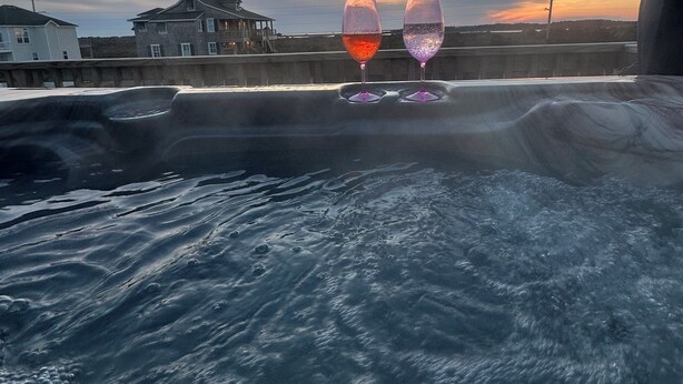 Uitzicht op het water met twee wijnglazen op een leuning, een zonsondergang op de achtergrond en een huis aan de overkant van het water.
