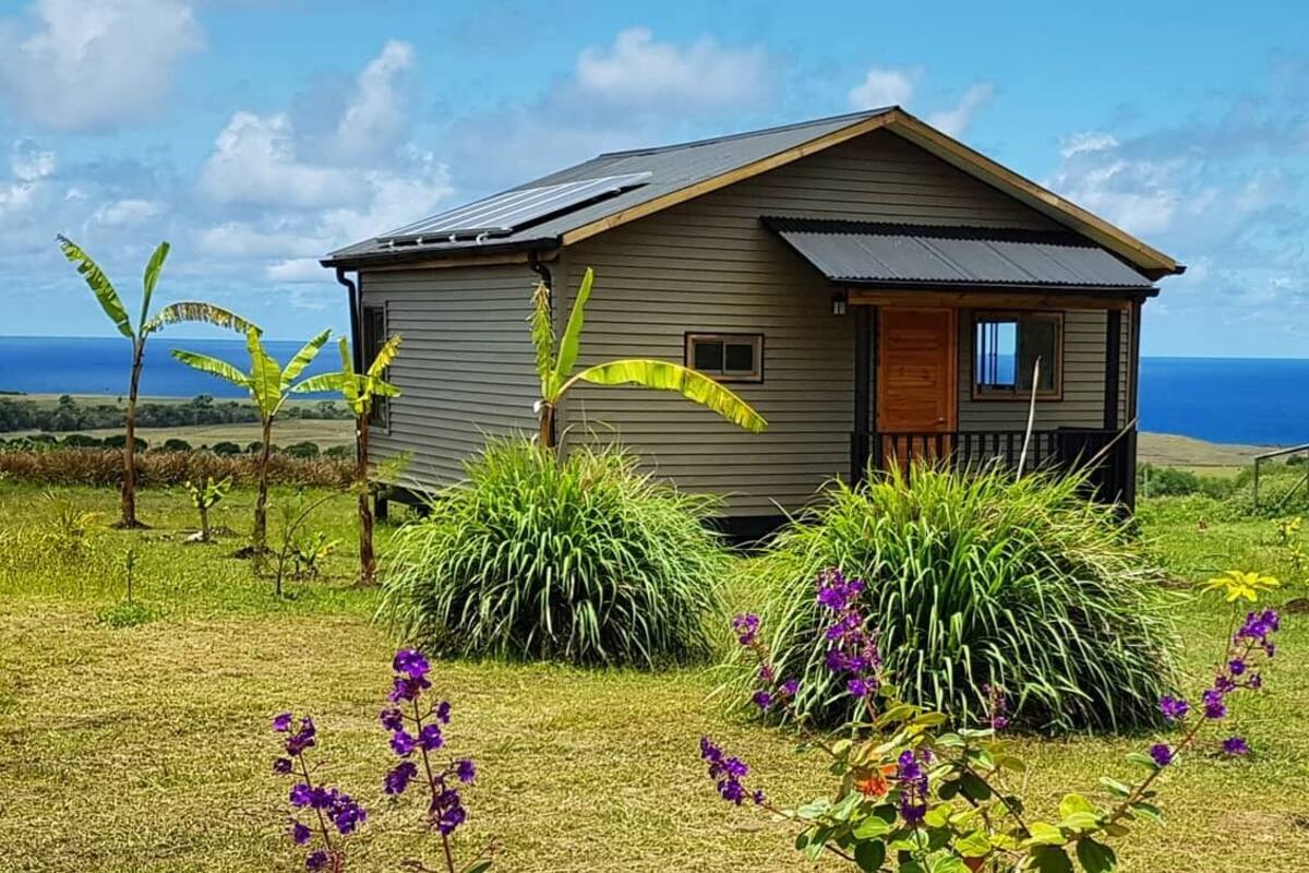 A small, single-story house with a sloped roof, solar panels, and a view of the ocean.