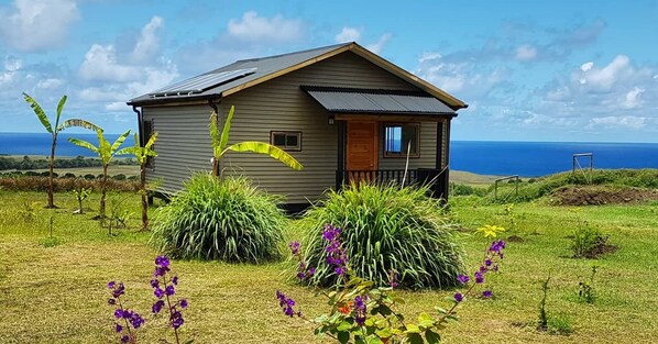 A small, single-story house with a sloped roof, solar panels, and a view of the ocean.