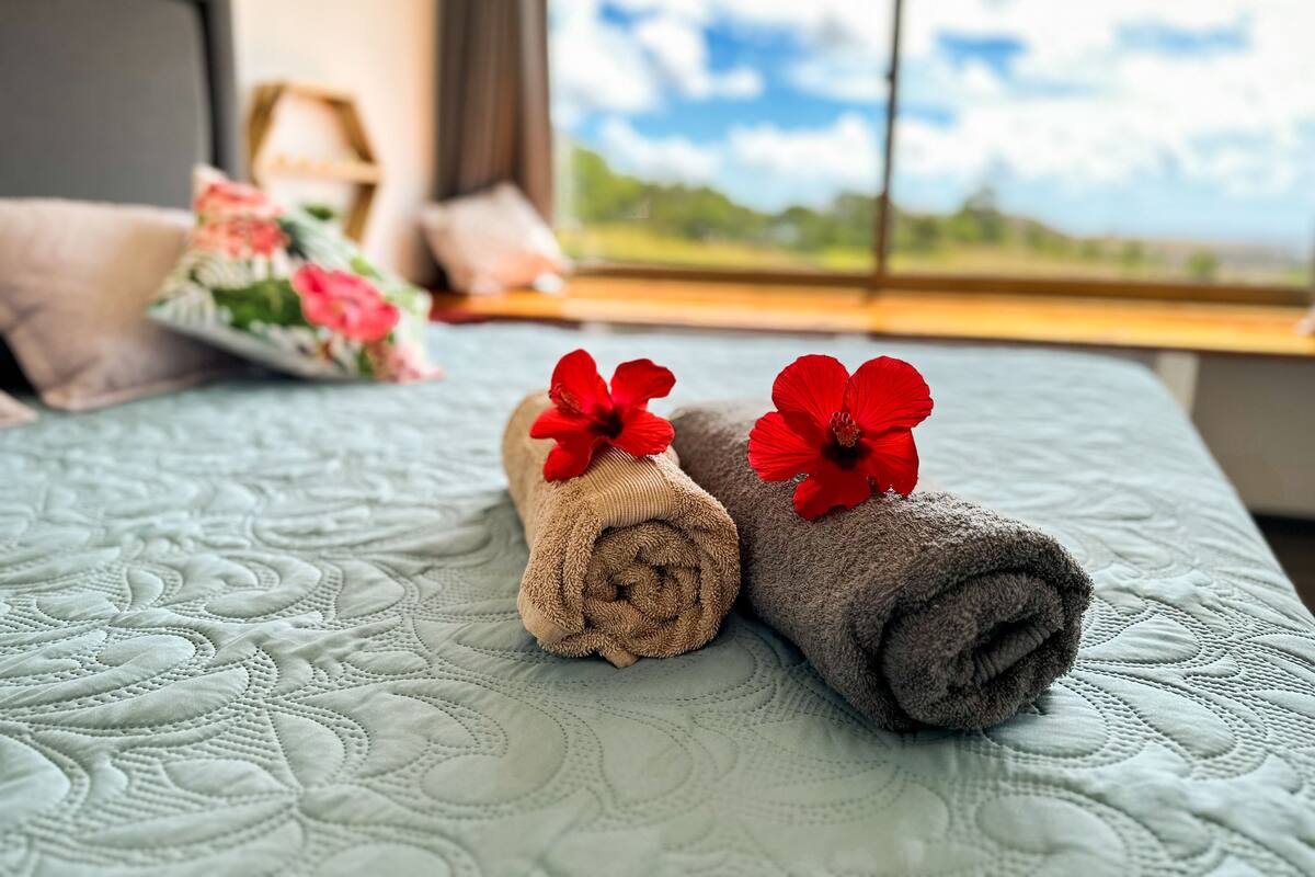 A bed with rolled towels and red flowers, a window with a view, and a bedside table with a lamp and decorative pillows.