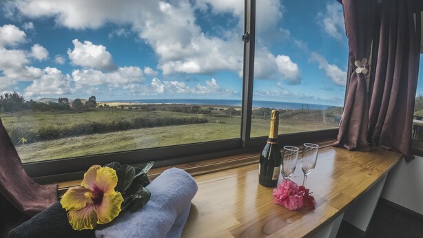 Une pièce dotée d’une grande fenêtre donnant sur un champ verdoyant et la mer. Sur le rebord de la fenêtre, on trouve une bouteille de champagne, deux verres, une serviette et un bouquet de fleurs.