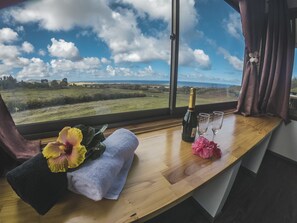 A room with a large window offering a view of a grassy field and the sea. On the windowsill, there is a bottle of champagne, two glasses, a towel, and a flower arrangement. - Maunga roa eco lodge (Hanga Roa)