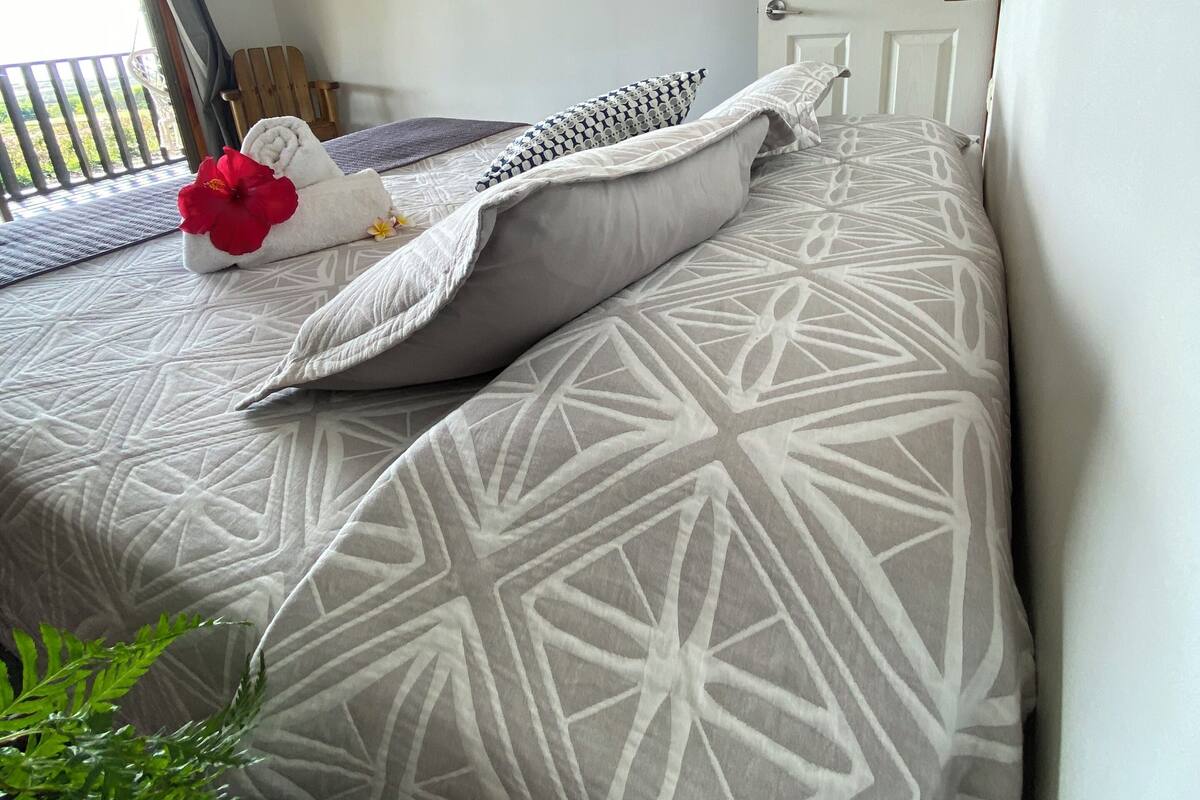 A neatly made bed with patterned bedding, a red flower, and a small potted plant.