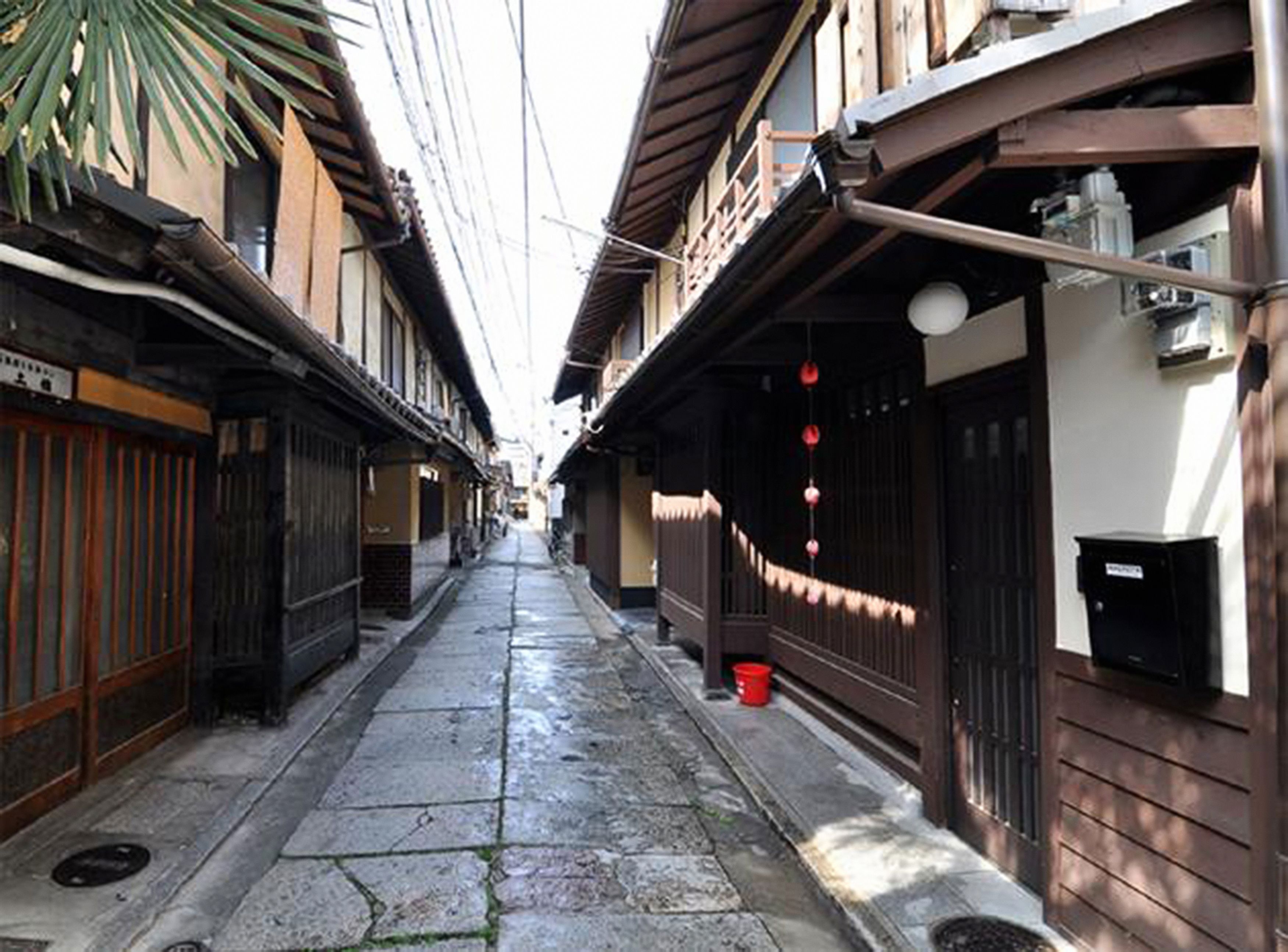 A narrow alleyway with traditional wooden buildings, a stone-paved path, and a red bucket on the ground.