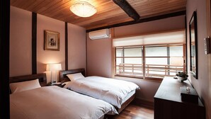 A hotel room with two beds, a wooden ceiling, an air conditioning unit, a window with blinds, and a bedside table with a lamp. - Kiyomizu Machiya Inn (Kyoto)