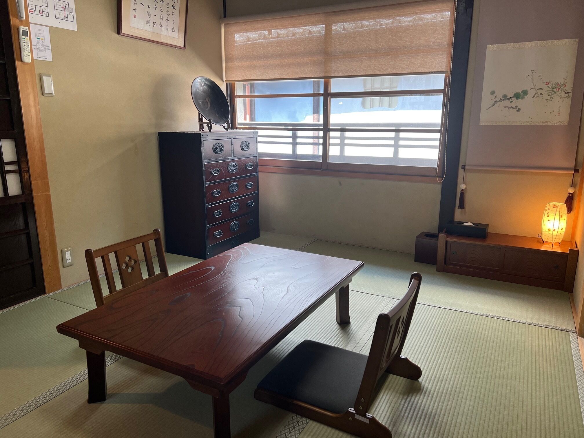 A traditional Japanese room with tatami flooring, a wooden table, chairs, a cabinet with drawers, a wall-mounted radio, and a window with a view.